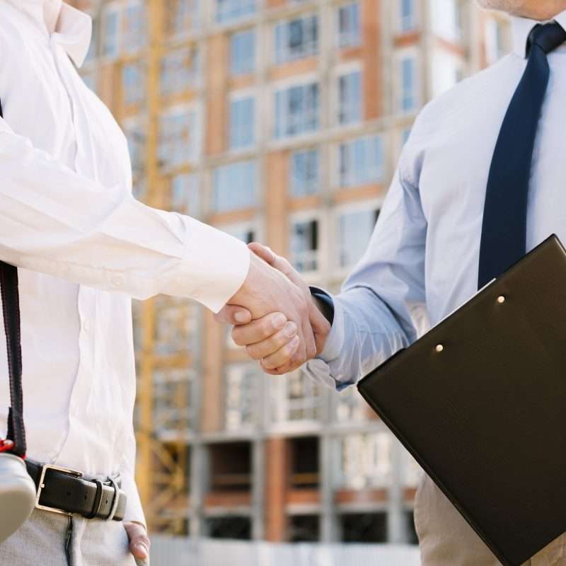 Two professionals shaking hands in front of a construction site, with one holding a clipboard, symbolizing a successful agreement