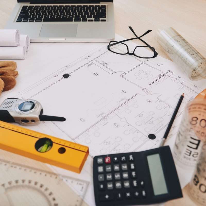 Construction blueprints, a laptop, calculator, and safety equipment laid out on a table