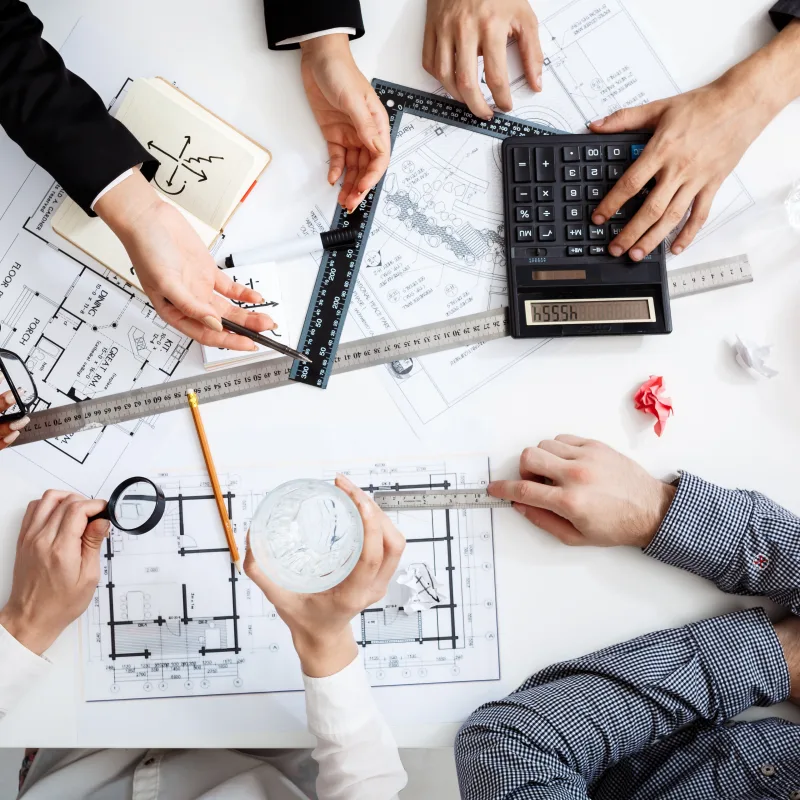 A top view of a diverse group of construction professionals collaborating over blueprints and project plans on a white table, with tools like rulers, calculators, and a hard hat.