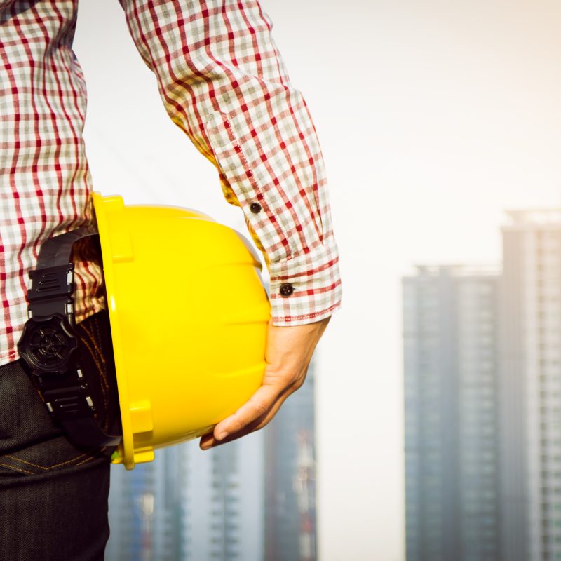Construction worker holding a yellow safety helmet with a background of modern high-rise buildings.