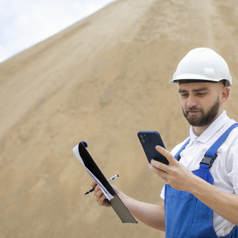 Construction worker using a mobile app on a smartphone while holding documents on a construction site.